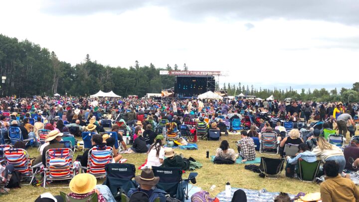 A large crowd of people all sit on the ground, facing a stage that says Winnipeg Folk Festival 